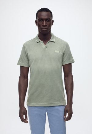 Man die recht vooruit kijkt, gekleed in een lichtgroen polohemd met korte mouwen en lichtblauwe broek, staand tegen een effen achtergrond.