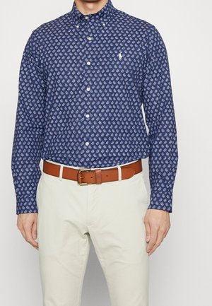 Homme portant une chemise bleue à motif cachemire boutonnée, un pantalon beige et une ceinture en cuir marron avec une boucle dorée.