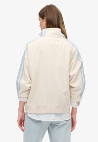 Sudadera crema con cuello alto, que presenta franjas laterales azules y un ajuste relajado. La tela texturada y los puños acanalados completan el diseño.