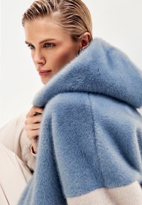 Cappotto di pelliccia azzurro chiaro con cappuccio, caratterizzato da una texture morbida. Il design presenta un motivo a blocchi di colore con accenti di beige chiaro.