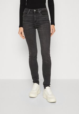 Kvinde iført sorte højtaljede skinny jeans, sort langærmet top og hvide plateau sneakers, der står mod en ensfarvet baggrund.