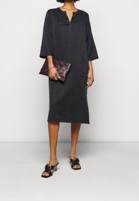 Musta polvipituinen mekko, jossa on V-pääntie ja kolmen neljänneksen hihat, sileä pinta. Pitää käsissään kudottua burgundinpunaa clutchia, yhdistettynä mustiin korkokenkiin, joissa on bows.