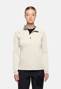 Jonge vrouw met blond haar draagt een lichtbeige pullover met kwartrits en zwarte broek, staand tegen een effen lichte achtergrond, recht vooruit kijkend.