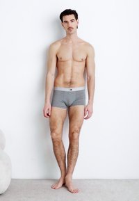 Boxers en coton gris avec une taille élastique arborant une marque. Le modèle, de constitution mince, pose devant un mur blanc.