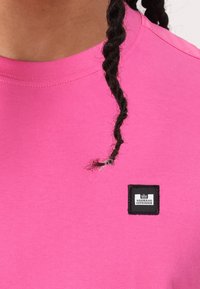 Camiseta de algodón rosa con escote redondo y una etiqueta cuadrada negra con texto blanco "WEEKEND OFFENDER" cosida en la parte delantera.