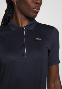 Polo shirt blu navy con colletto, tre bottoni e logo verde, rosso e bianco sul lato sinistro del petto. Texture del tessuto liscia.