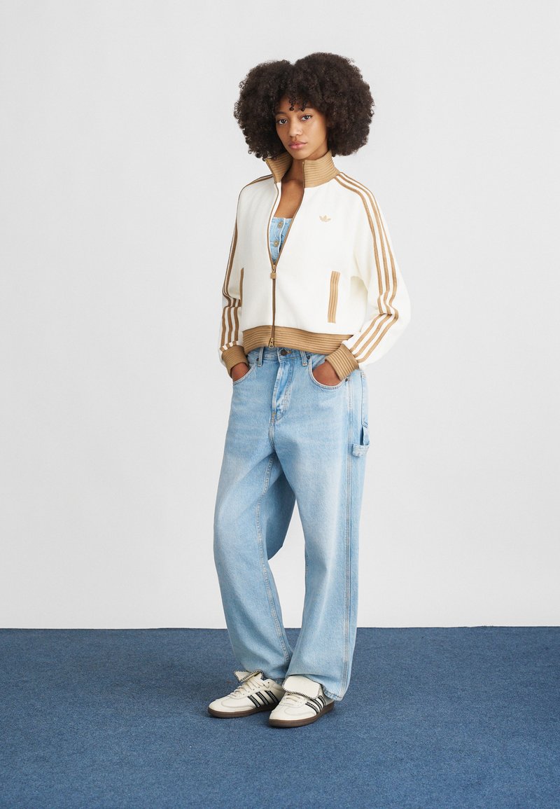 Veste zippée blanche et marron avec col côtelé et trois rayures marron. Jean baggy bleu clair. Chaussures blanches avec des accents noirs.