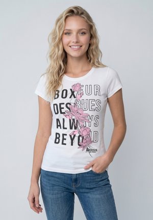 Junge Frau mit welligem blondem Haar lächelt, trägt ein weißes T-Shirt mit schwarzem Text und rosa Blumendesign, kombiniert mit blauen Jeans.