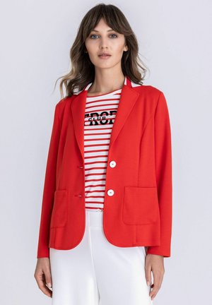 Frau mit welligem Haar, die ein rotes Sakko, ein weißes und rot gestreiftes Hemd mit Text und eine weiße Hose trägt, steht vor einem schlichten hellen Hintergrund.