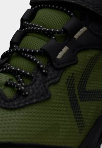 Chaussure de sport verte avec une tige texturée, des accents noirs et des lacets à motifs. Elle comprend un verrou de lacet et des zones renforcées pour plus de durabilité.