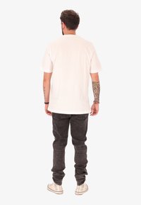 Uomo con tatuaggi su entrambe le avambraccia che indossa una maglietta bianca semplice, jeans scuri e scarpe da ginnastica alte bianche, in piedi di spalle.