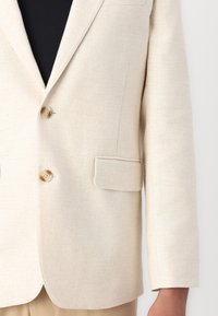 Lichtbeige blazer, gestructureerde stof, twee knopen en een enkele borstzak. Gepuncteerde revers en driekwartmouwen.