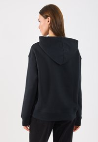 Zwarte hoodie met een ruime capuchon, relaxte pasvorm, en geribbelde manchetten en zoom. Zachte stof; gedragen met losse zwarte broek.