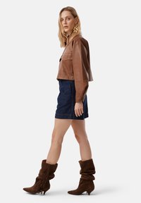 Chaqueta de cuero marrón corta, pantalones cortos de mezclilla oscura y botas de ante marrón con detalle de flecos. Modelo de perfil contra un fondo blanco.