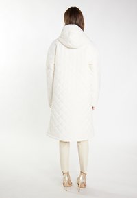 Gequiltede witte jas met een capuchon, met een lange, verlengde vorm en een gladde textuur. Gecombineerd met lichtgekleurde broek en hakken.