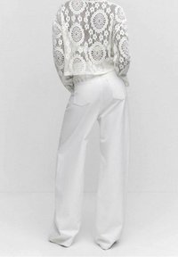 Osoba ubrana w białe szerokie spodnie i białą koronkową bluzkę z długim rękawem, stojąca na jednolitym jasnym tle, plecami zwrócona do widza.