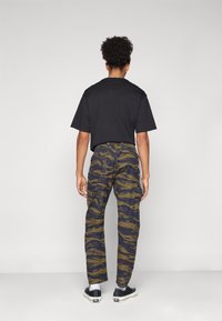 Persona de pie de espaldas, vistiendo camiseta negra, pantalones cargo verdes y negros con diseño, calcetines blancos y zapatillas negras.