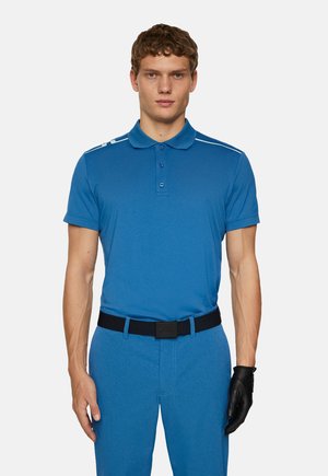 Polo bleu en tissu lisse avec un col à trois boutons, des accents aux épaules, associé à un pantalon assorti et une ceinture noire.