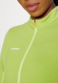 Gros plan d'une personne portant un haut zippé Mammut vert clair avec col montant et tirette de zip orange.