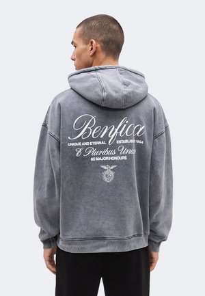 Joven de espaldas que lleva una sudadera con capucha gris con "Benfica" y texto relacionado impreso en la parte trasera, de pie frente a un fondo liso.