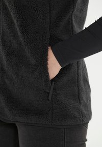 Mustavilla fleece-vesti, jossa on pehmeä ja tekstuurinen pinta. Varustettu sivutaskulla, jossa on vetoketju, ja pitkät mustat hihat alla.