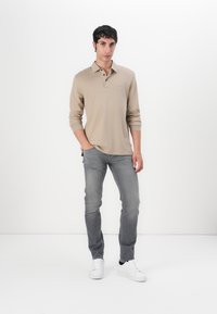 Beige long-sleeve polo shirt with three buttons, paired with slim gray jeans and white sneakers, set against a plain white background.