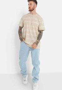 Beige t-skjorte med hvit kursiv tekstmønster, sammen med lyseblå jeans og hvite joggesko. Modellen har tatoveringer og middels langt bølgete hår.