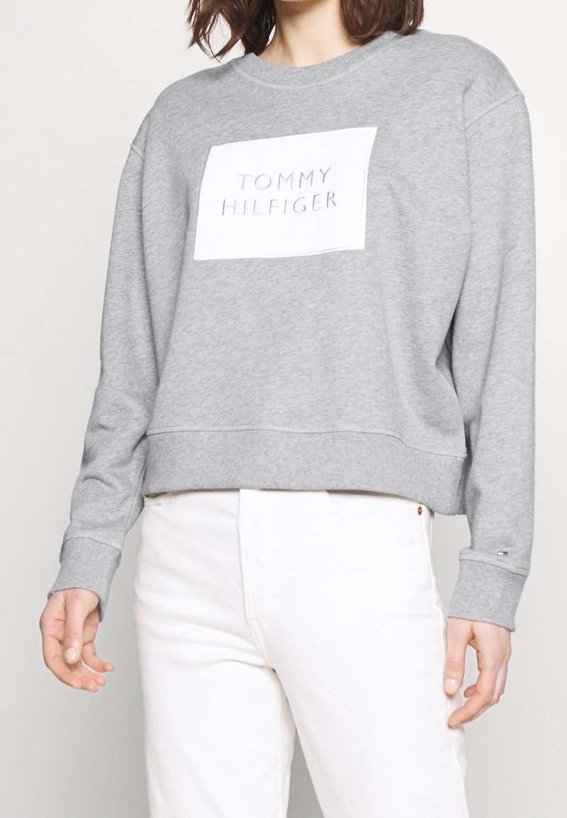 Gray sweatshirt with a crew neckline and long sleeves. Features a central white patch with "TOMMY HILFIGER" in bold letters.
