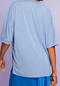 Persona con una blusa corta de manga corta de color azul claro y pantalones azules brillantes, con una pulsera de plata en la muñeca, de pie contra un fondo púrpura.