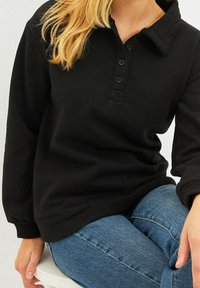 Suéter negro de estilo poncho con un cuello tipo camisa, tres botones delanteros y puños acanalados. Combinado con jeans azules. Textura de tela suave.