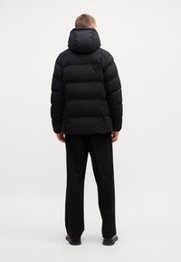Jachetă puffer neagră cu glugă, având un design matlasat și o textură lucioasă. Afișată pe un model din spate, asortată cu pantaloni negri.