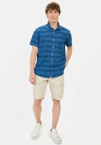 Blå kortærmet skjorte med knapper og horisontalt stribemønster samt lomme, parret med beige cargo-shorts og hvide sneakers.