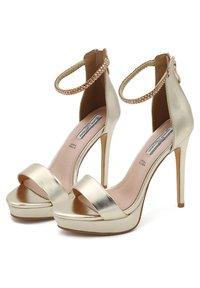 Sandali con tacco alto in metallo dorato, caratterizzati da una ampia fascia anteriore, una cinghietta alla caviglia con dettagli intrecciati e una piattaforma per un'altezza extra.