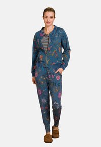 Blauwe jas met bloemenpatroon en een ritssluiting, gecombineerd met bijpassende joggingsbroeken. Kenmerken zijn ribgebreide manchetten en tailleband. Model draagt bruine slippers.