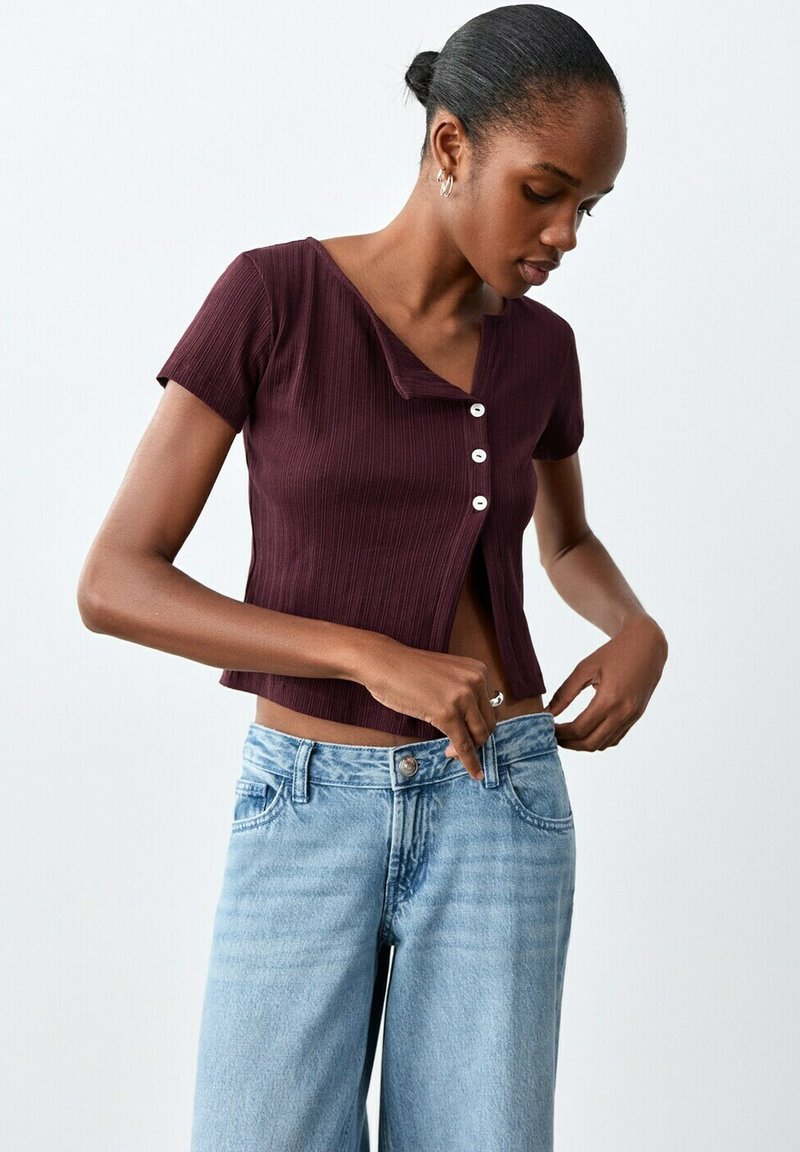 Femme aux cheveux foncés coiffés en chignon, portant un haut boutonné manches courtes couleur bordeaux et un jean taille haute bleu clair, ajustant la taille de son jean.