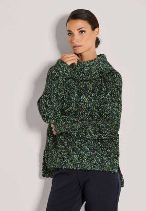 Frau mit dunkelhaarigen, zurückgebundenen Haaren, die einen grob gestrickten Pullover mit grünen, blauen und schwarzen Flecken trägt und vor einer hellen Wand steht.