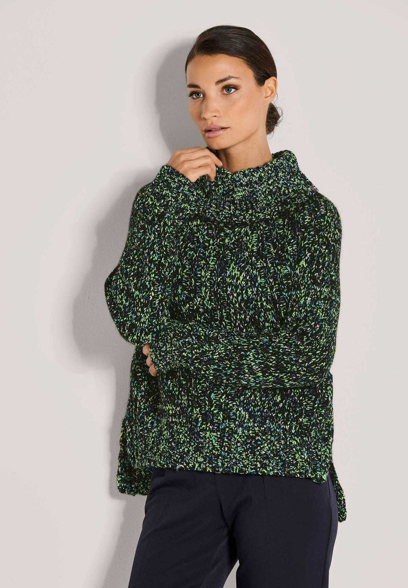 Frau mit dunkelhaarigen, zurückgebundenen Haaren, die einen grob gestrickten Pullover mit grünen, blauen und schwarzen Flecken trägt und vor einer hellen Wand steht.