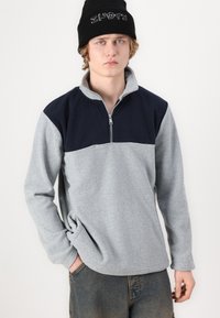 Pullover in felpa grigio e blu navy con colletto a mezza zip, superficie testurizzata e maniche lunghe. Accompagnato da un berretto di maglia nera.