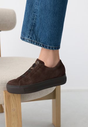 Pied portant une basket en daim marron foncé avec semelle noire reposant sur une chaise beige rembourrée, jean bleu raccourci au-dessus de la cheville.