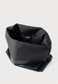 Sac matelassé noir avec une base en similicuir noir texturé et une étiquette. Il présente une ouverture supérieure souple et volumineuse pour un accès facile.