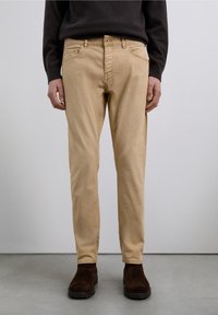 Beige chinos i bomuld med en lige pasform, der har forlommer og lynlås, kombineret med mørkebrune støvler i ruskind. Glat tekstur.
