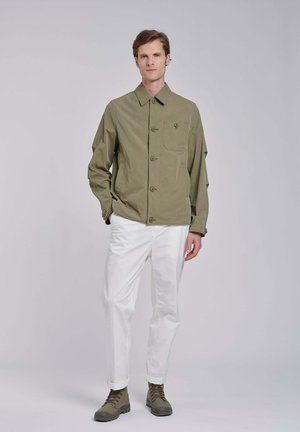 Mężczyzna w oliwkowo-zielonej kurtce zapinanej na guziki, białych spodniach i brązowych butach stoi na tle jednolitego, jasnego tła.