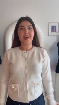 Νεαρή γυναίκα με μακριά σκούρα μαλλιά που φορεί λευκή ζακέτα με κουμπιά, στέκεται μέσα σε ένα δωμάτιο με μια κορνίζα και μια κούκλα μοντελισμού πίσω της.