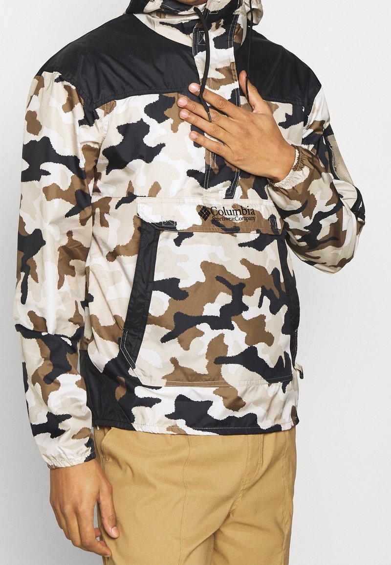 Veste camouflage présentant un panneau d'épaule noir, design à demi-fermeture éclair, poche avant et poignets élastiques ; couleurs comprenant beige, marron et noir.