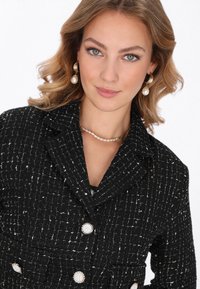 Blazer in tweed nero con una finitura testurizzata, caratterizzato da accenti dorati, grandi bottoni e un colletto. Completato con gioielli di perle.