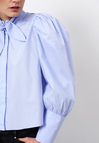 Light blue blouse featuring a bow tie at the neck, smocked detailing at the shoulders, and voluminous puff sleeves.