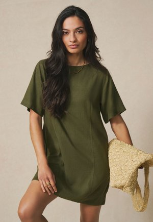 Woman with long dark hair wearing an olive green short-sleeve dress holding a woven straw bag against a neutral background.