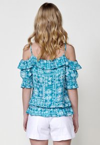 Top turchese a spalle scoperte con maniche a volant, decorato con motivi floreali e geometrici bianchi, abbinato a shorts bianchi.