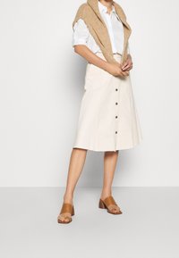 Lys beige nederdel med knaplukning foran, knælang, lavet af tekstureret stof. Sammen med en hvid skjorte og sandfarvede åbne tå blokhæle.