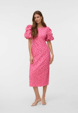 Kvinde stående iført en lyserød midi kjole med puffede ærmer og blomsterprint, kombineret med hvide sandaler med hæl og stropper.